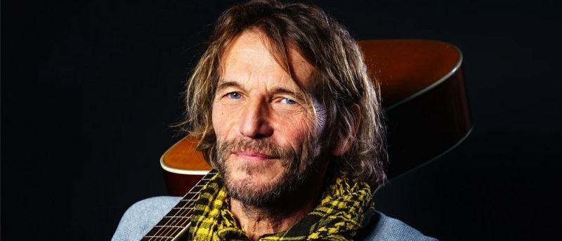 Ein Mann mit langen Haaren und Bart in einem blauen Hemd und gelbem kariertem Schal, der eine Gitarre mit einem lächelnden Gesicht vor schwarzem Hintergrund hält.