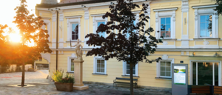 A yellow building with a red roof and windows. A statue and a tree are in front. A bench and a plant are nearby.