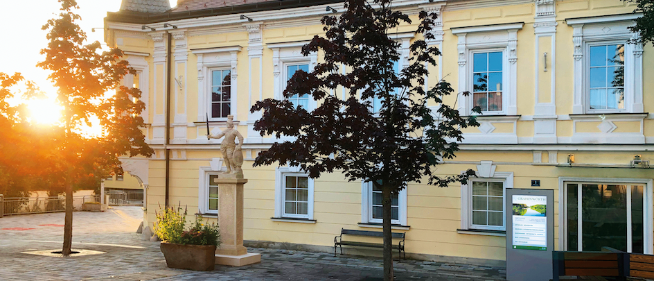 Ein gelbes Gebäude mit einem roten Dach und Fenstern. Eine Statue und ein Baum stehen davor. Eine Bank und eine Pflanze sind in der Nähe.