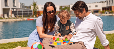Eine Familie genießt einen Tag am See. Die Eltern und ihr Kind sitzen auf einer Decke auf dem Gras. Das Kind spielt mit Spielzeug.