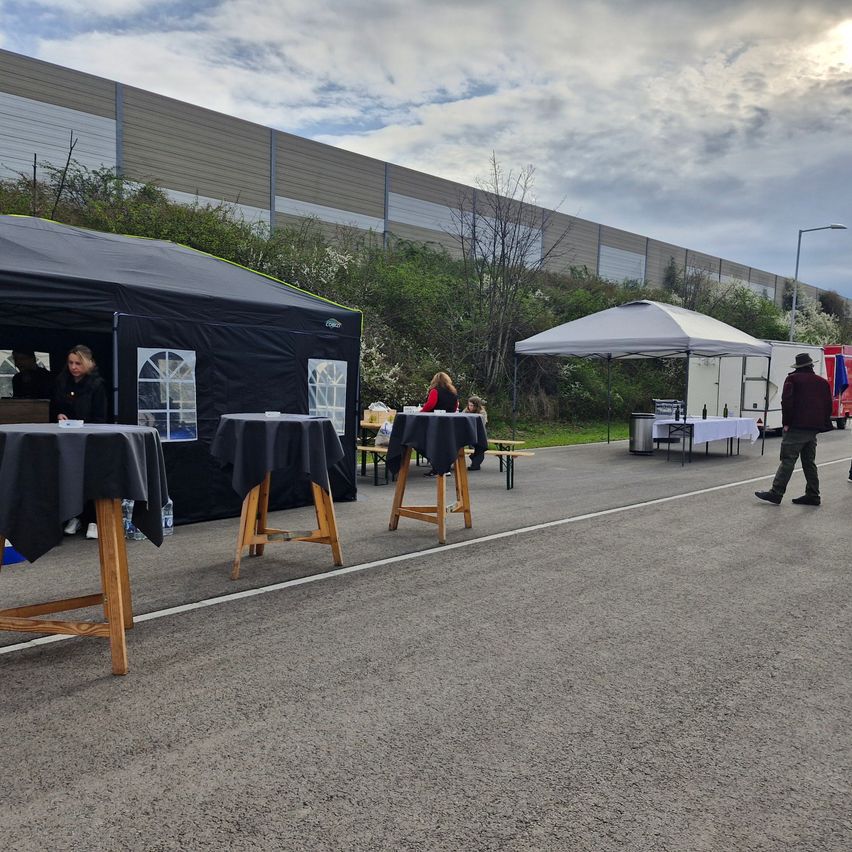 Two tents are set up in an open area, one black and one white. A man walks past, and people sit inside the tents. Tables are set up outside with bottles and chairs.