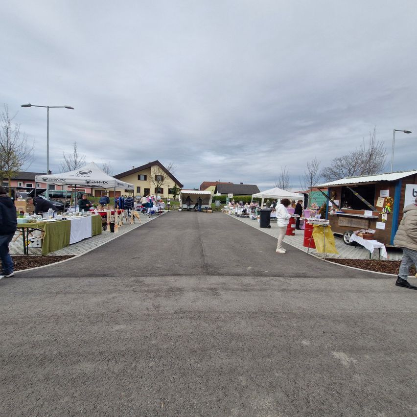 An outdoor market with tents and stalls. People are walking and shopping. Buildings and trees are in the background.