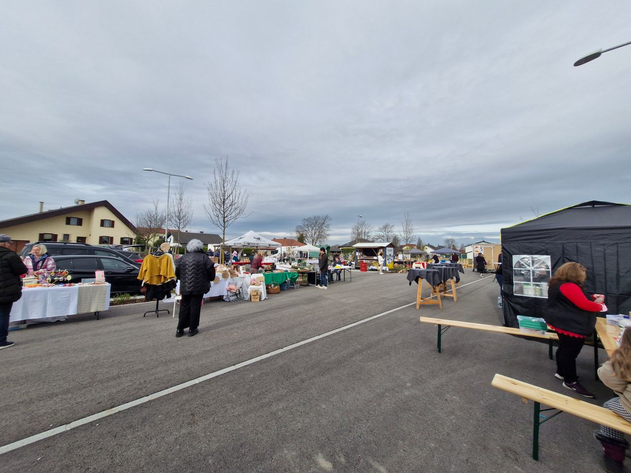 A flea market is set up on a street. Several stalls are seen selling various items. A woman stands near a table. Trees and houses are in the background.