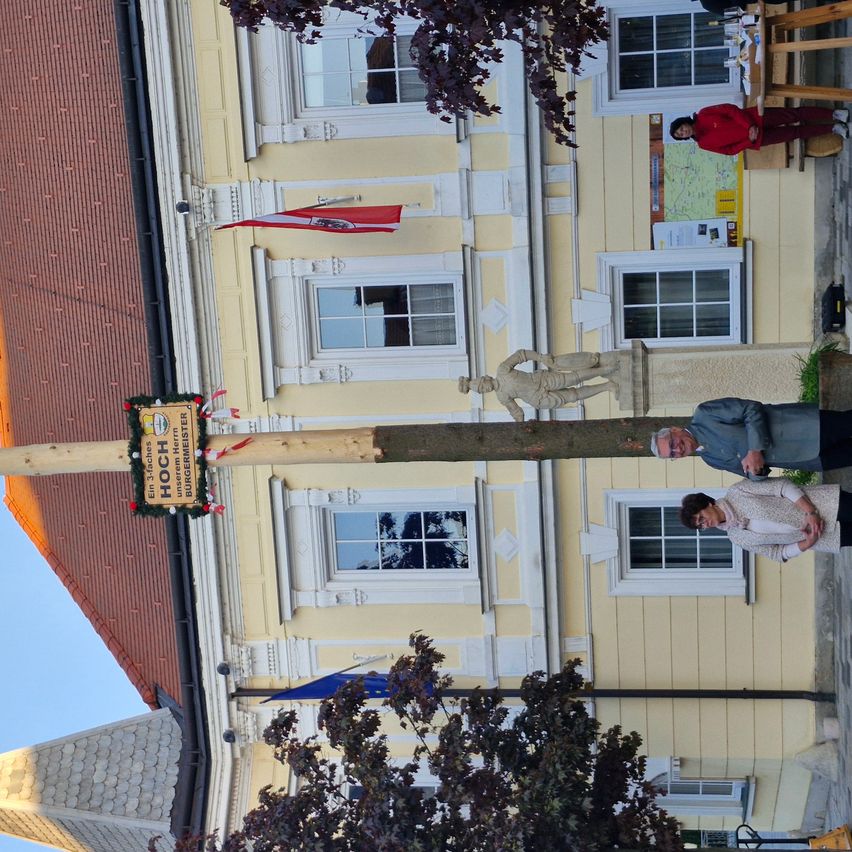 An aerial view shows a yellow building with many windows. A pole with a sign and a flag stands in front. Two people are standing beside a tree.