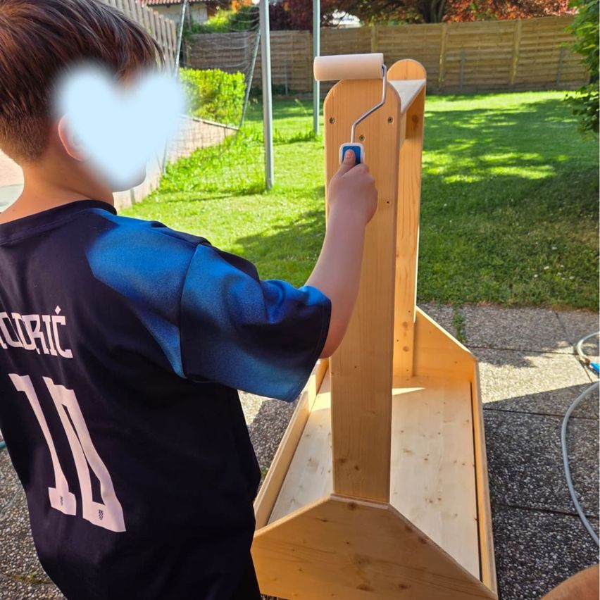 Ein Junge in einem blauen Shirt mit der Nummer 14 darauf malt eine Holzstruktur. Er hält einen Malerwalze mit einem weißen Roller auf einem Holzständer.