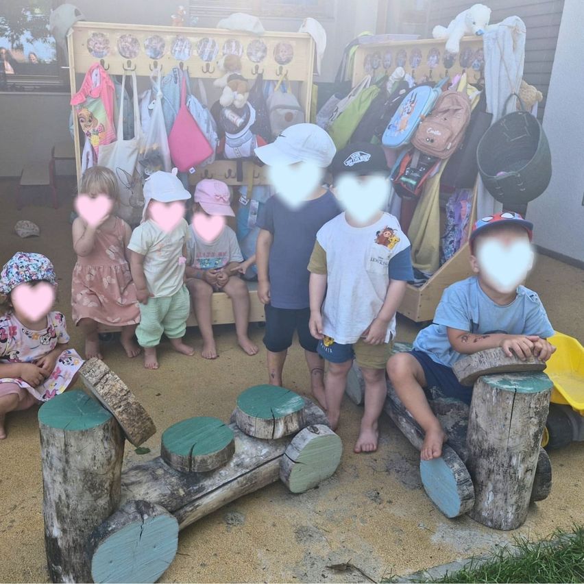 Mehrere Kinder sitzen auf Holzstümpfen, einige stehen. Sie tragen Hüte und einige eine Brille. Dahinter befinden sich Regale mit Taschen, und einige hängen an der Wand.