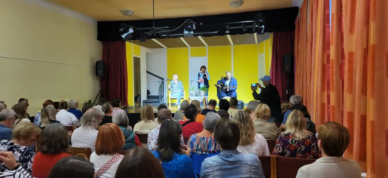 Drei Personen mit Musikinstrumenten sind auf der Bühne in einem Theater. Die beiden Frauen sitzen und der Mann steht. Ein Publikum sitzt vor ihnen.