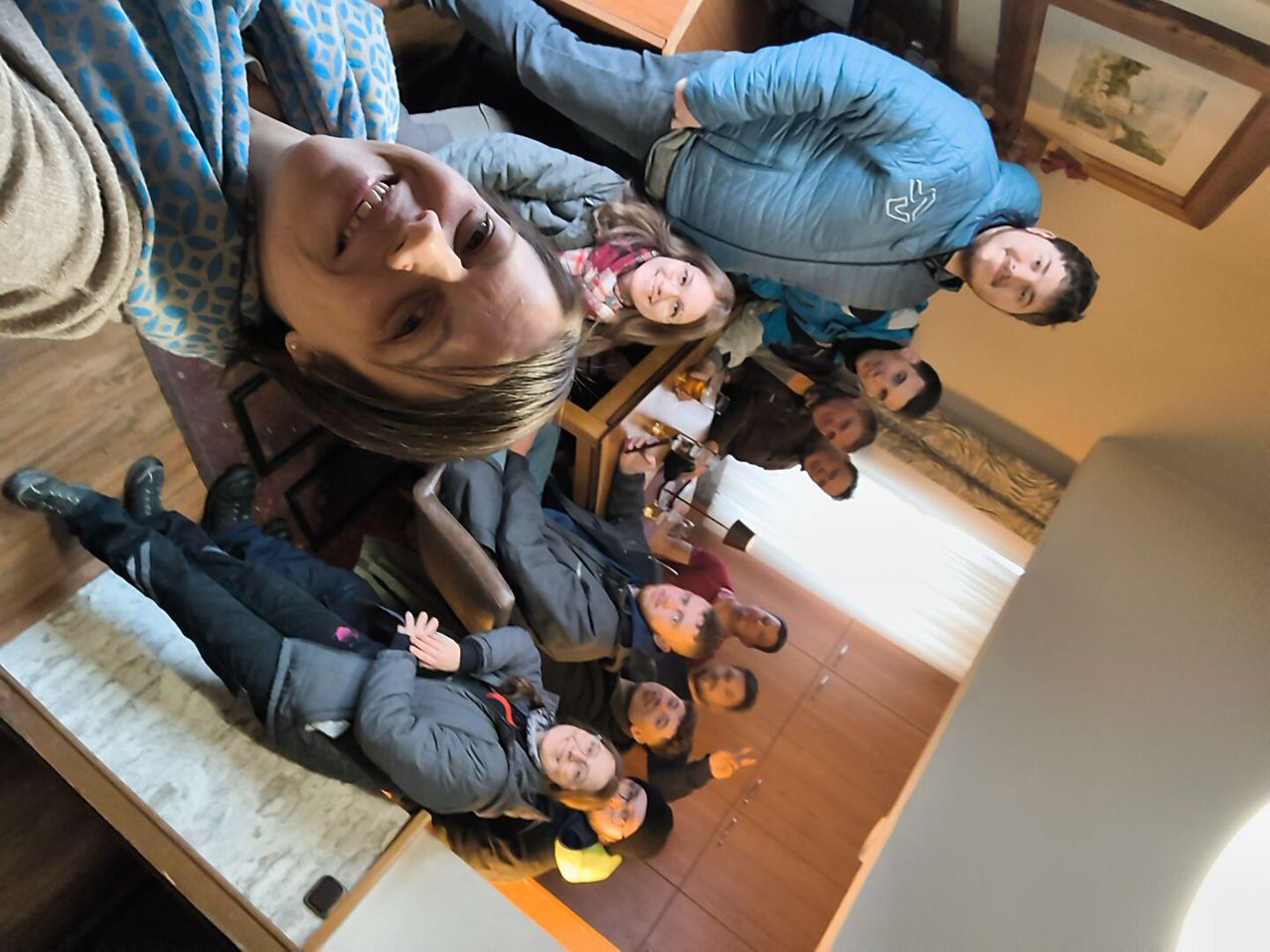 Eine Gruppe von Menschen ist drinnen versammelt, lächelt und posiert für ein Foto. Einige sitzen auf Stühlen, andere stehen. Eine Person trägt eine blaue Jacke mit einem weißen Logo. Der Raum hat Holzböden und weiße Wände.