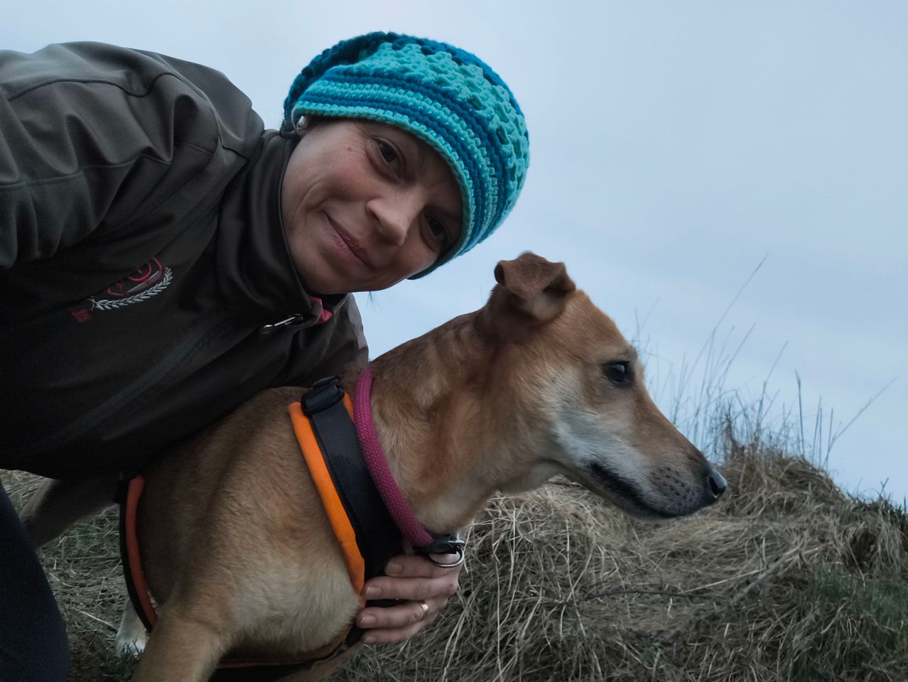 A woman with a blue beanie poses with her brown dog, both smiling. The dog wears a reflective orange and pink collar. They stand on dry grass.