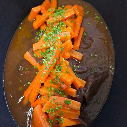 Eine Schüssel gefüllt mit einer reichhaltigen braunen Suppe, belegt mit Scheiben von orangefarbenen Karotten und grünen Kräutern, die auf einer schwarzen Oberfläche ruht.