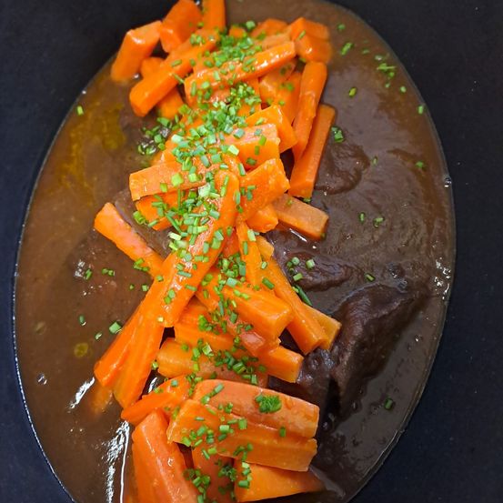 Eine Schüssel gefüllt mit einer reichhaltigen braunen Suppe, belegt mit Scheiben von orangefarbenen Karotten und grünen Kräutern, die auf einer schwarzen Oberfläche ruht.