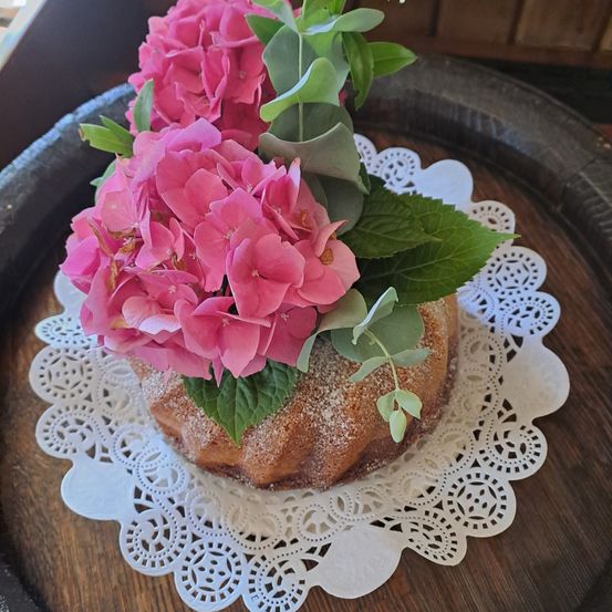 Ein Kuchen mit rosa Blumen darauf steht auf einem weißen Doily auf einem Holztisch. Der Kuchen ist mit Zucker bestreut.