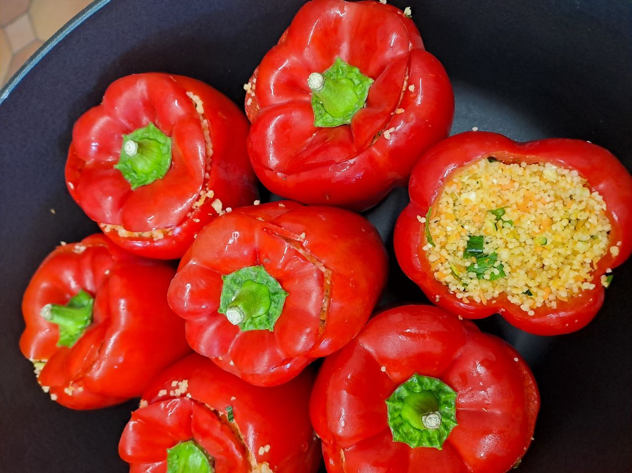 Eine Nahaufnahme mehrerer roter Paprikaschoten, von denen einige ihren Deckel entfernt haben und eine Füllung aus gelbem Couscous und grünen Kräutern zeigen.