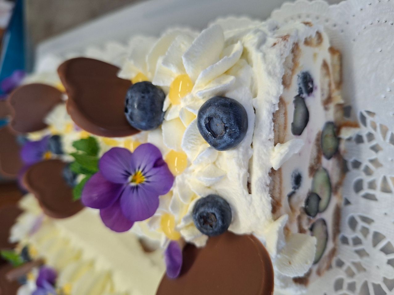 Ein Nahaufnahme von einem Kuchen mit Schlagsahne, Blaubeeren und Schokoladenverzierungen. Eine lila Blume befindet sich auf der linken Seite.