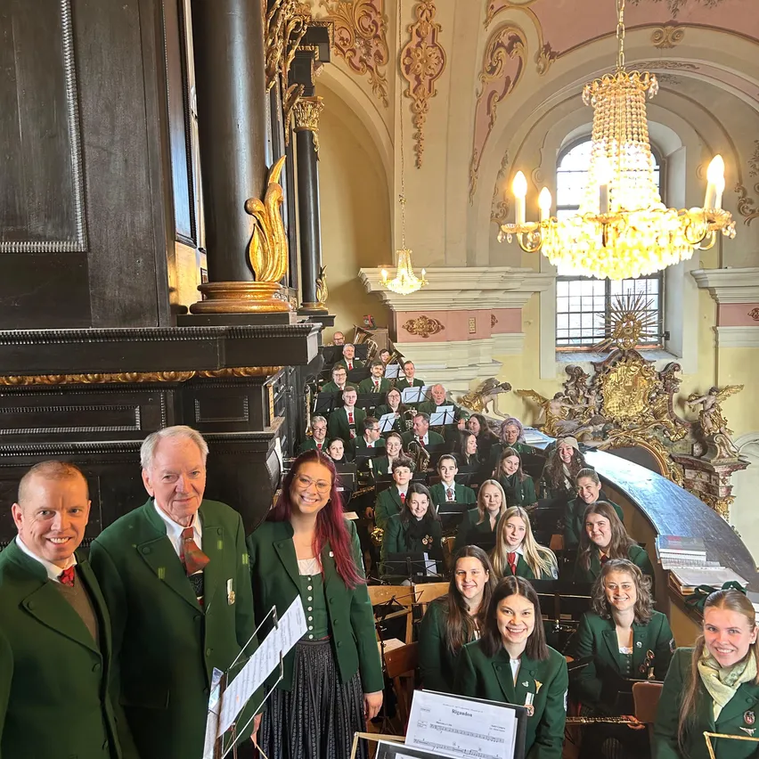 Eine Gruppe von Musikern in grünen Uniformen, die für ein Foto in einem historischen Gebäude mit verzierten Säulen, einem Kronleuchter und Musikinstrumenten posiert.