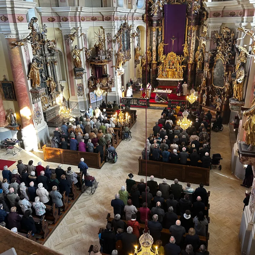 Eine große Gemeinde nimmt in einer Kathedrale mit goldenen Altären und Statuen an einem Gottesdienst teil. Die Menschen sitzen auf hölzernen Kirchenbänken, und zwei Priester stehen am Altar.