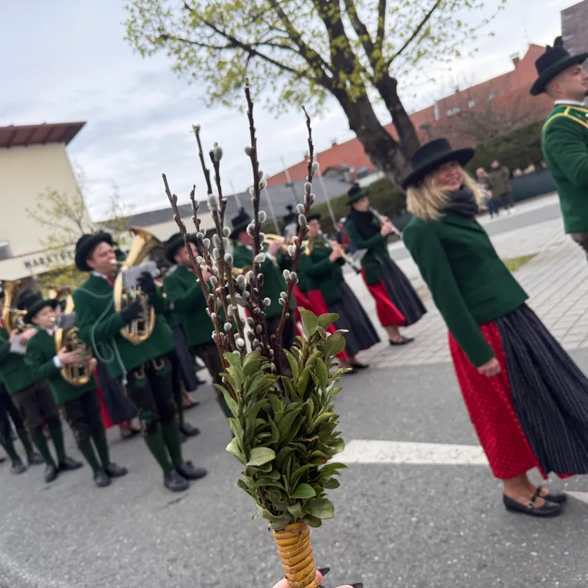 Eine Person mit einem Blumenstrauß steht vor einer Blaskapelle in traditioneller Kleidung, die auf einer Stadtstraße auftritt.