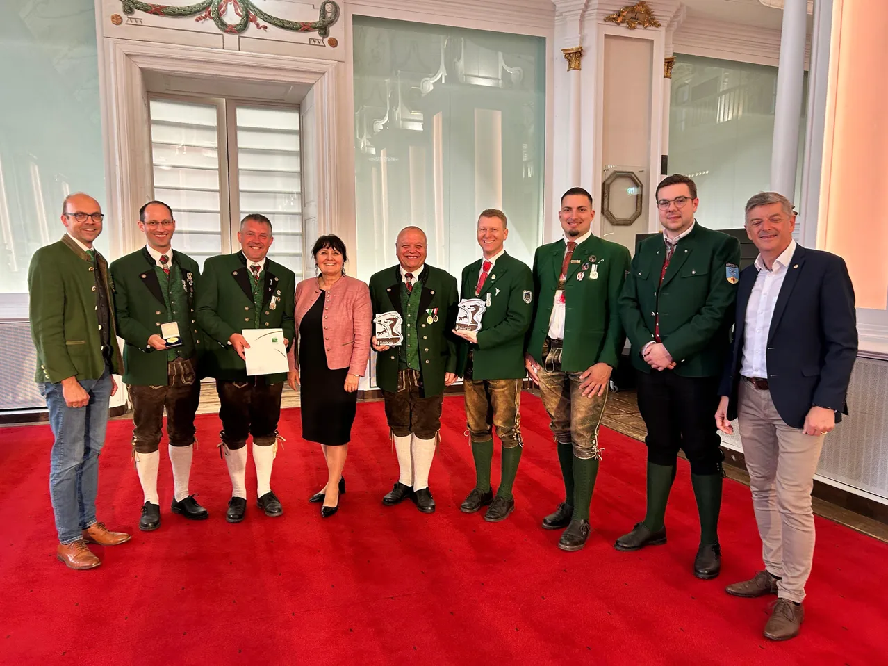 Eine Gruppe von Männern und einer Frau in traditioneller bayerischer Tracht stehen auf einem roten Teppich, halten Auszeichnungen und lächeln. Sie befinden sich in einem großen Saal.