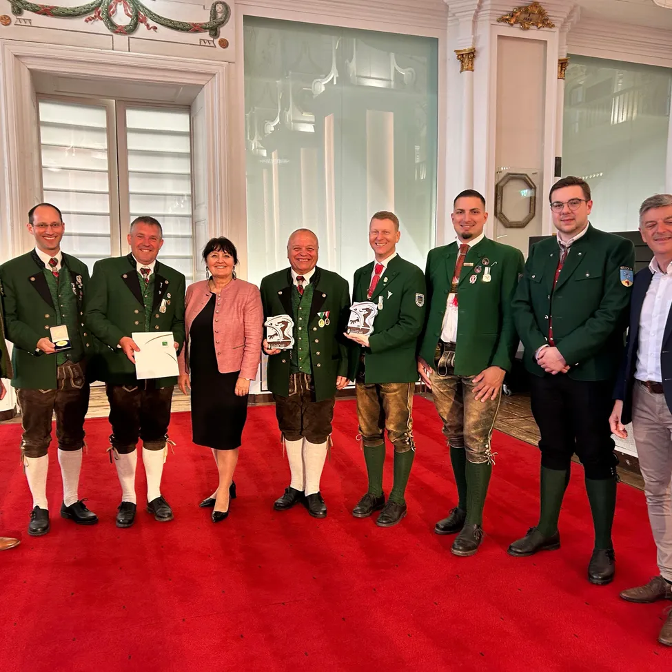 Eine Gruppe von Männern und einer Frau in traditioneller bayerischer Tracht stehen auf einem roten Teppich, halten Auszeichnungen und lächeln. Sie befinden sich in einem großen Saal.
