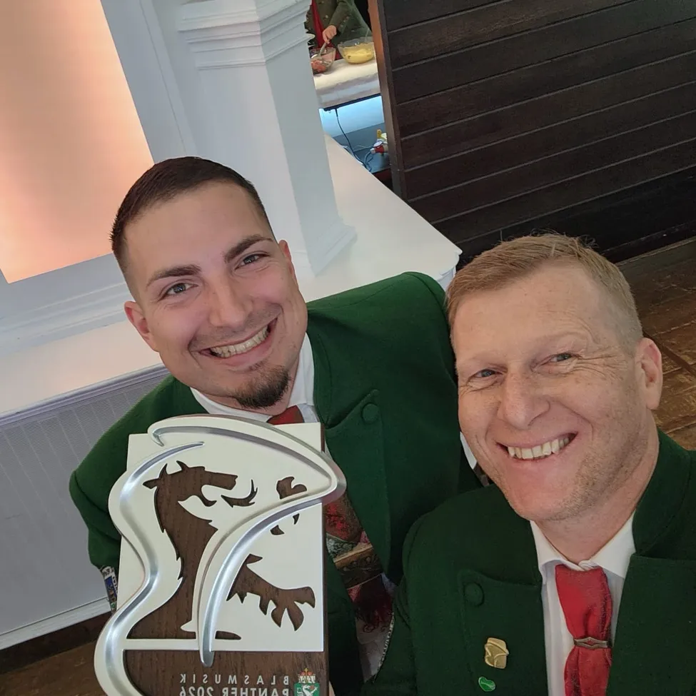 Zwei Männer in grünen Uniformen, einer hält eine Trophäe mit einem Drachen und lächelt in einem Raum.
