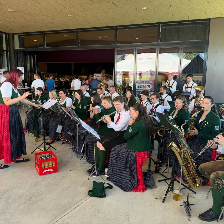 Eine Gruppe von Musikern, in grünen und roten Kostümen, tritt vor einem Gebäude mit großen Fenstern auf. Eine Frau in einem roten Kleid steht neben einem roten Kasten.