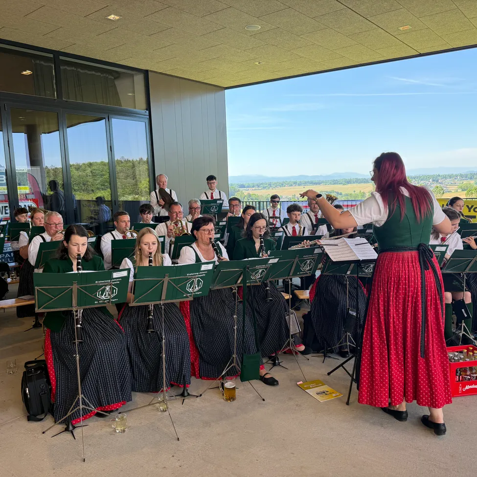 Eine Gruppe von Musikern in traditioneller grüner und roter Kleidung spielt in einem Raum mit großen Fenstern. Die Frau vor ihnen dirigiert, während sie steht.