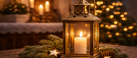 Eine Kerzenlaterne mit brennender Kerze steht auf einem Holztisch, umgeben von Tannenzweigen und Sternen, mit einem Weihnachtsbaum im Hintergrund.