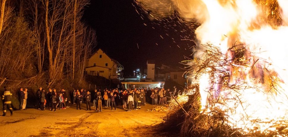 Eine Menschenmenge versammelt sich um ein großes Lagerfeuer in der Nacht. Das Feuer brennt hell und Bäume umgeben das Gebiet.
