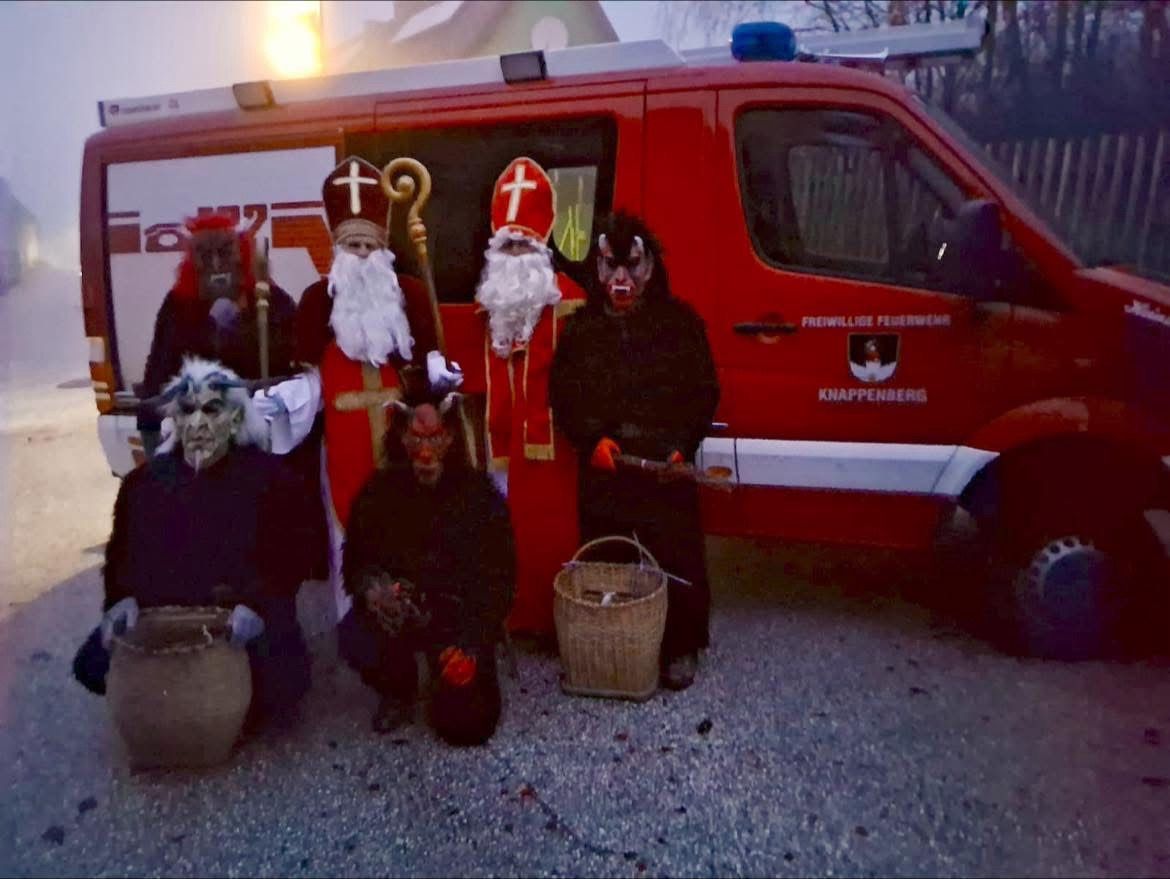 Eine Gruppe von Menschen in Kostümen posiert vor einem roten Feuerwehrauto mit der Aufschrift 'Freiwillige Feuerwehr Knappenberg.' Einige von ihnen tragen Masken.
