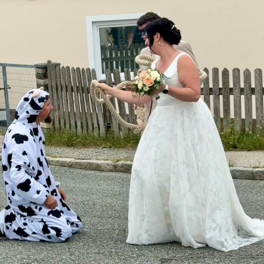 Ein Mann in einer Kuh-Verkleidung kniet auf der Straße und hält ein Seil, während eine Frau in einem Hochzeitskleid in der Nähe steht.