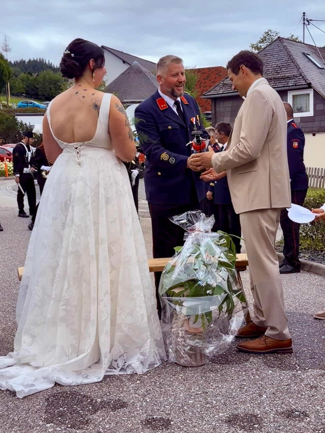 Eine Hochzeitszeremonie mit einer Braut in einem weißen Kleid, einem Bräutigam in einem hellbraunen Anzug und einem Mann in Uniform, der eine Pflanze in einer Plastiktüte präsentiert.