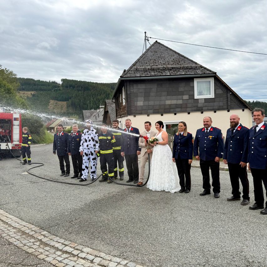 Eine Hochzeitsgesellschaft steht vor einem Haus, wobei Feuerwehrleute im Hintergrund Wasser sprühen.