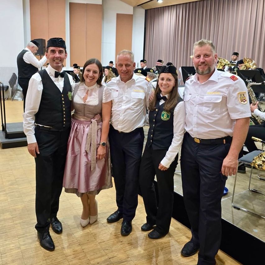 Vier Personen stehen zusammen in einem Raum mit einer Band im Hintergrund. Zwei Männer und zwei Frauen, alle in Uniform.