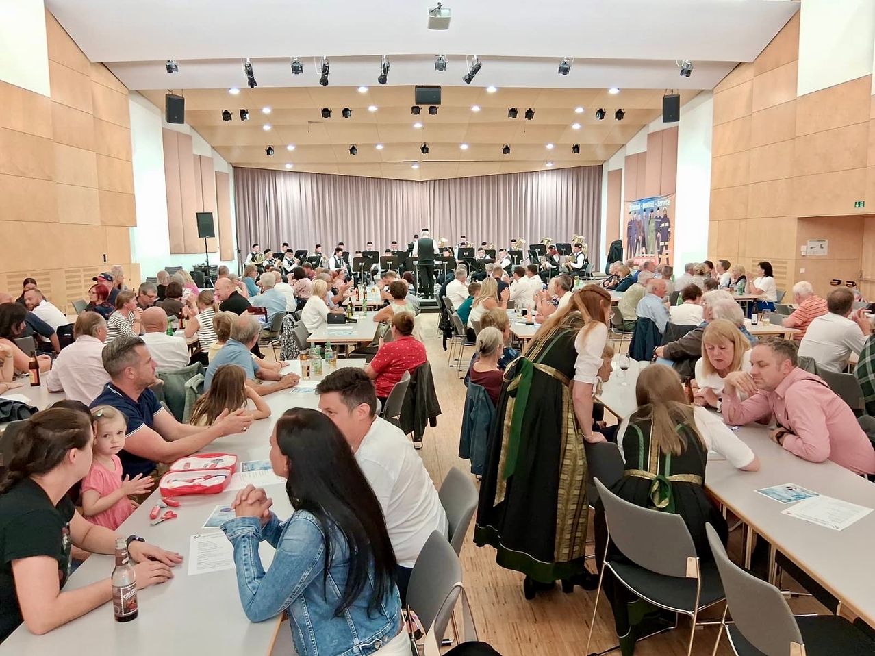 Eine große Indoor-Veranstaltung mit einer Band auf der Bühne, Gästen an Tischen, einige in traditioneller Kleidung, mit Holzboden und Bühnenhintergrund.