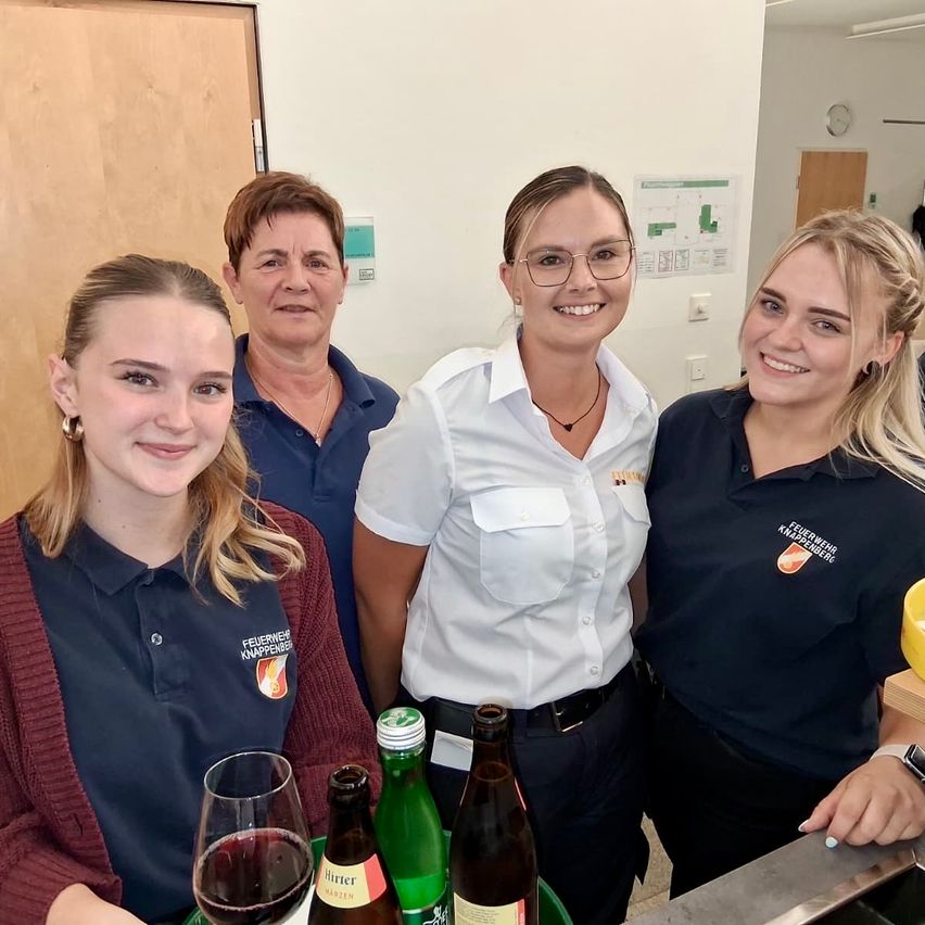 Vier Frauen in Uniform, zwei mit Brille, lächeln und posieren für ein Foto mit Getränken und einer Uhr im Hintergrund.