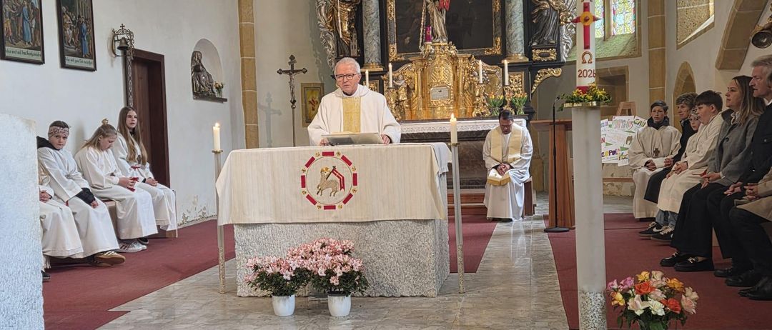 Ein Priester steht am Altar und liest aus einem Buch, mit rosa und weißen Blumen auf beiden Seiten. Hinter ihm schmücken Gemälde und Kerzen den Altar.