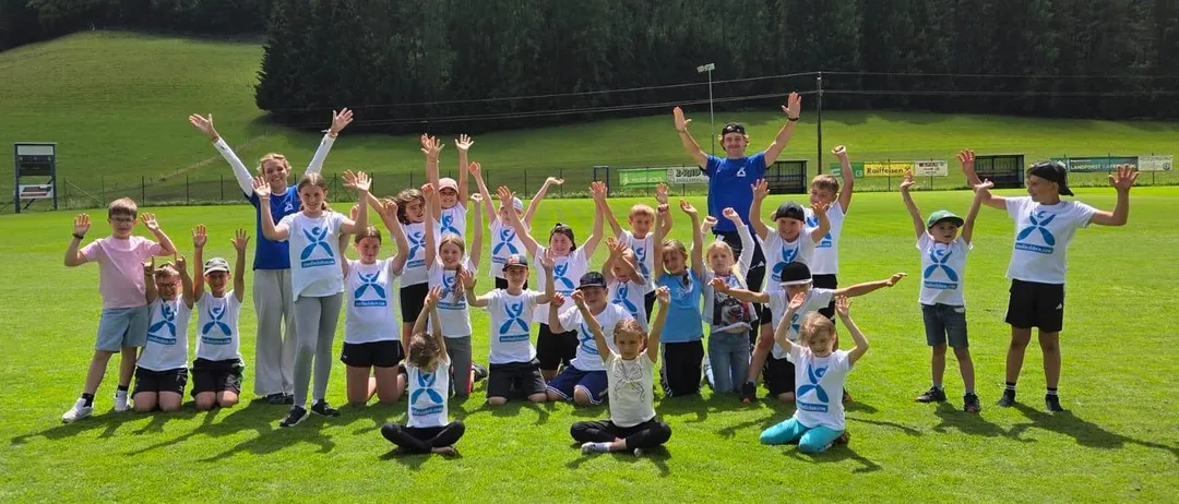 Eine Gruppe von Kindern und zwei Erwachsenen posiert für ein Foto auf einem Rasenfeld mit Bergen im Hintergrund.