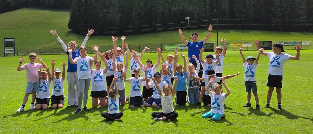 Eine Gruppe von Kindern und zwei Erwachsenen posiert für ein Foto auf einem Rasenfeld mit Bergen im Hintergrund.