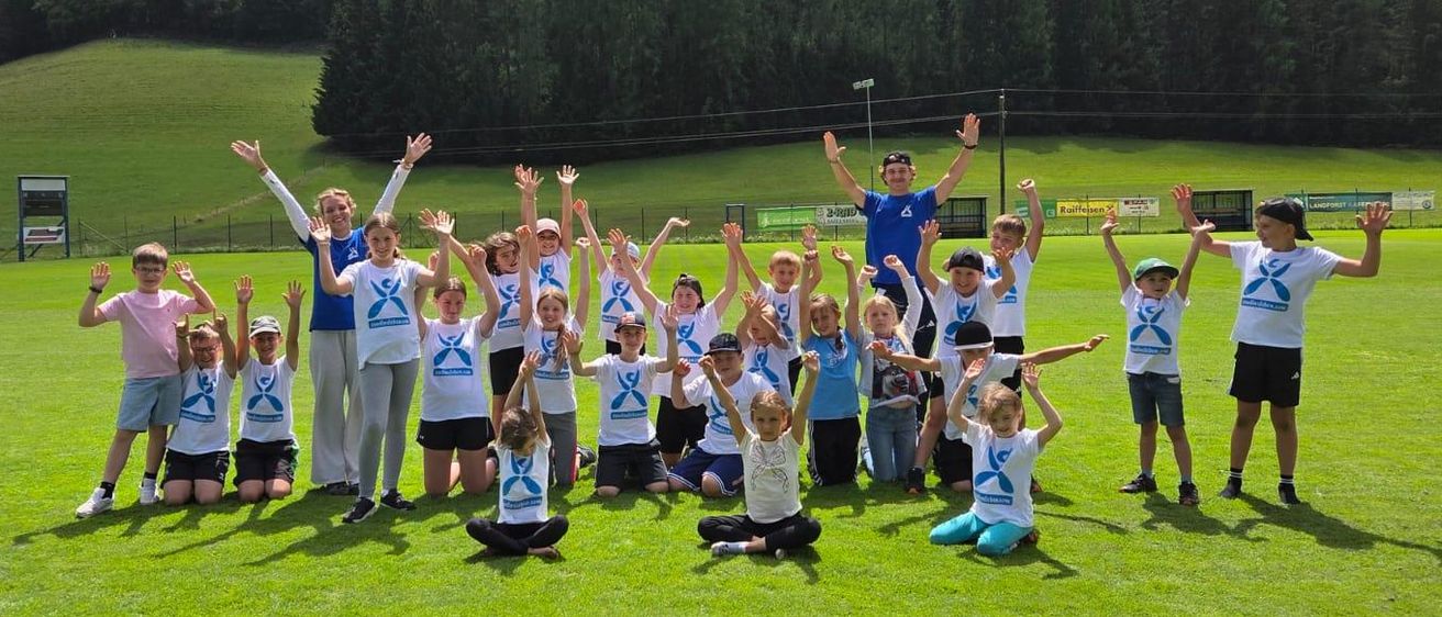 Eine Gruppe von Kindern und zwei Erwachsenen posiert für ein Foto auf einem Rasenfeld mit Bergen im Hintergrund.