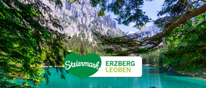 Ein ruhiger See mit einem Bergrücken im Hintergrund, mit einem grünen Herzlogo, das 'Steiermark' und einen weißen Banner mit 'ERZBERG LEOBEN' zeigt.