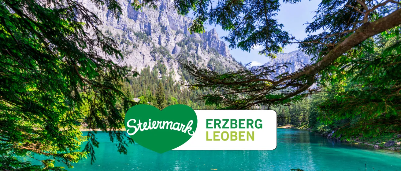 Ein ruhiger See mit einem Bergrücken im Hintergrund, mit einem grünen Herzlogo, das 'Steiermark' und einen weißen Banner mit 'ERZBERG LEOBEN' zeigt.