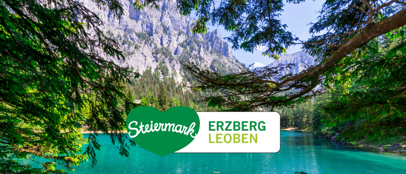 Ein ruhiger See mit einem Bergrücken im Hintergrund, mit einem grünen Herzlogo, das 'Steiermark' und einen weißen Banner mit 'ERZBERG LEOBEN' zeigt.