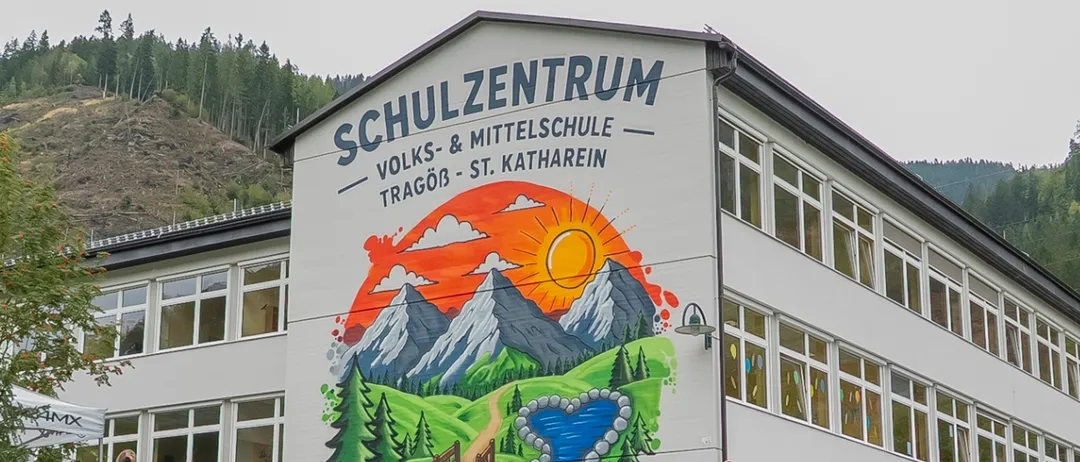 Ein großes Gebäude mit einem Wandbild von Bergen und einem Fluss ist zu sehen. Viele Menschen sind draußen versammelt, wahrscheinlich auf etwas wartend.