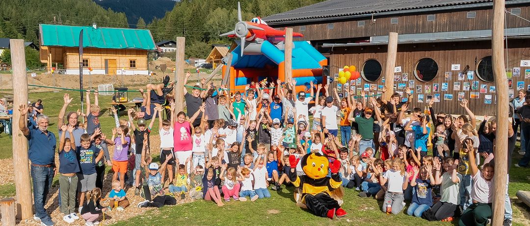 Eine große Gruppe von Kindern und Erwachsenen posiert für ein Foto vor einem Gebäude mit einem großen aufblasbaren Flugzeug obenauf.