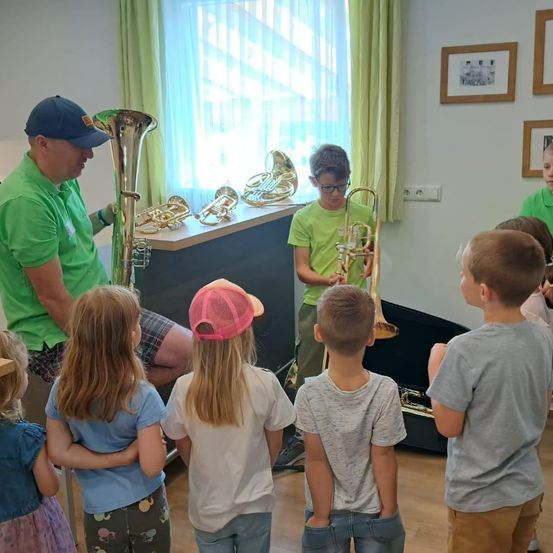 Ein Mann in einem grünen Shirt demonstriert ein Kind eine Posaune in einem Raum mit Fenster und Bilderrahmen.
