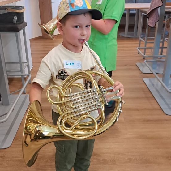Ein junger Junge namens Liam hält eine goldene französische Trompete in einem Raum. Er trägt einen Hut und ein T-Shirt mit einem Dinosaurier-Design. Hinter ihm ist eine weitere Person in einem grünen Shirt zu sehen.