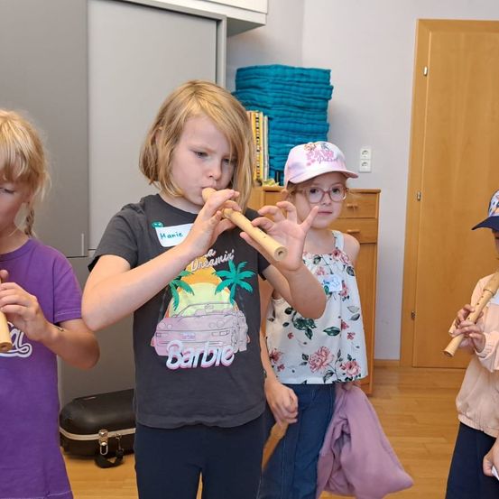 Vier Kinder stehen in einem Raum und spielen Holzflöten. Ein Mädchen in einer rosa Mütze und Brille spielt eine Flöte mit beiden Händen. Ein anderes Mädchen in einem lila Shirt spielt eine Flöte mit einer Hand. Dahinter sind ein Schrank mit Schubladen, ein Regal mit Büchern und ein Stapel blauer Decken zu sehen.