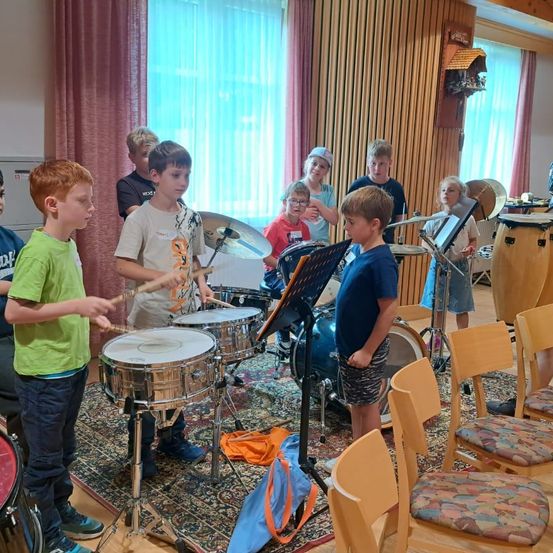 Eine Gruppe von Kindern spielt in einem Raum mit Holzwänden und Teppichtrommeln. Einige tragen Brillen, und ein Junge hält einen Drumstick. Musikinstrumente und Stühle sind um sie herum angeordnet.