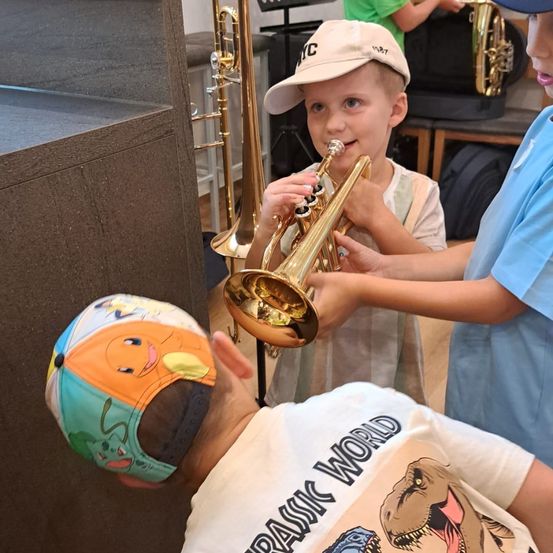 Zwei kleine Jungen spielen Trompeten in einem Musikgeschäft. Ein Junge trägt einen Hut und der andere eine Baseball-Kappe. Ein weiterer Junge lugt von hinten hervor.