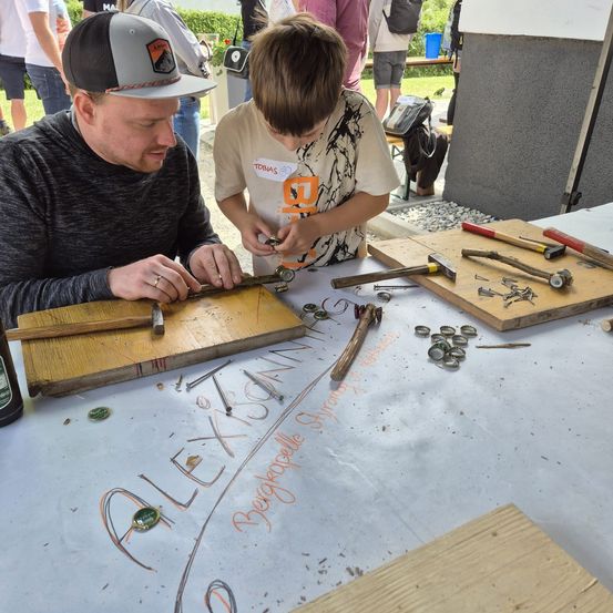 Ein Mann und ein Junge arbeiten mit Werkzeugen an einem Projekt auf einem Tisch. Sie tragen Freizeitkleidung. Der Junge trägt ein weißes T-Shirt mit orangefarbenem Aufdruck.